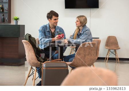 Two young adult couple in denim jackets chat at small round table in airport lounge with suitcase and bags nearby Two young adult couple in denim jackets chat at small round table in airport lounge with suitcase and bags nearby 133962813