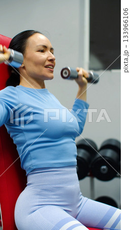 Deltoid workout with dumbbells - close-up of a fit Caucasian woman during gym workout. Deltoid workout with dumbbells. Physical education as a norm of life to maintain health. 133966106