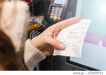 A woman reviews her shopping receipt at a self-service checkout in a store, ensuring her expenses align with her family budget during a busy shopping trip 133970751