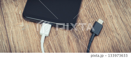 Black smartphone with charger cables on wooden background. Telephone charging concept Black smartphone with charger cables on wooden background. Telephone charging concept 133973633