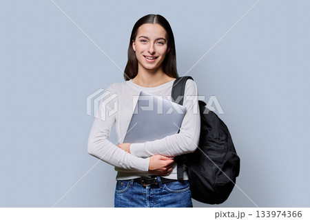 Portrait of beautiful female teenage student with backpack, laptop on gray background 133974366