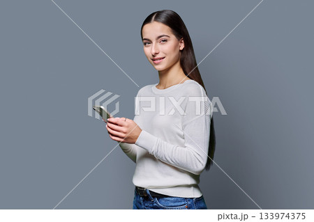 Portrait of smiling teenage girl holding smartphone on gray studio background Portrait of smiling teenage girl holding smartphone on gray studio background 133974375
