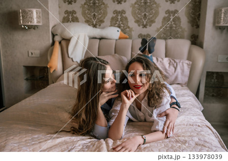 Women Bedroom Friendship Goose Pillow: Two friends whisper secrets, lying on a bed with a goose-shaped pillow, indoors. 133978039