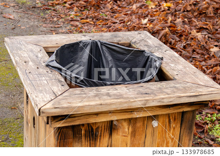 A wooden trash can with a black bag sits in a park area with fallen leaves around it. The scene shows late autumn conditions A wooden trash can with a black bag sits in a park area with fallen leaves around it. The scene shows late autumn conditions 133978685