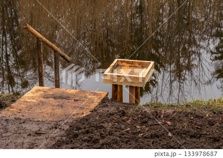 A wooden structure stands near a pond surrounded by trees. The platform connects to a box that is next to the water A wooden structure stands near a pond surrounded by trees. The platform connects to a box that is next to the water 133978687