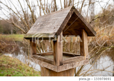 A wooden bird feeder is placed on a post next to a pond surrounded by bare branches and autumn scenery A wooden bird feeder is placed on a post next to a pond surrounded by bare branches and autumn scenery 133978691