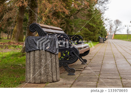 Two benches and a covered trash bin sit along a path in a park surrounded by trees on a cloudy day in the early afternoon 133978693