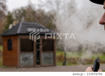 A person smokes from an electronic device while standing near a small coffee kiosk outside during a cloudy day 133978700