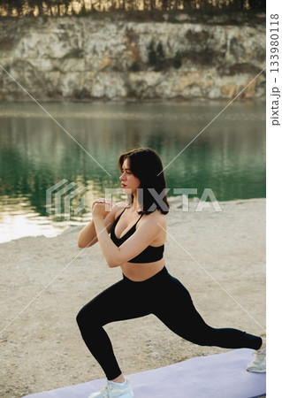 Woman practicing lunges by a serene lake during a sunny day with scenic cliffs in the background 133980118