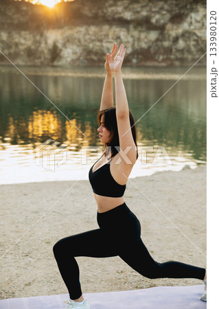 Woman practicing yoga on the shore of a peaceful lake during sunset surrounded by nature and calm water 133980120