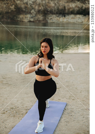 Woman practicing yoga on a mat by the serene water at sunset, showcasing strength and balance in a beautiful natural setting 133980121