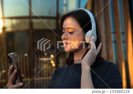 Young woman listens to music with headphones while using a smartphone in a bright urban setting during sunset Young woman listens to music with headphones while using a smartphone in a bright urban setting during sunset 133980129