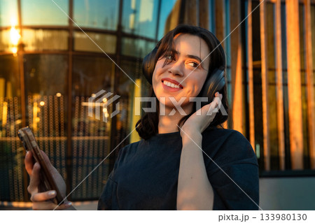 Young woman enjoys music while listening on headphones at sunset near a modern building in a vibrant urban setting 133980130