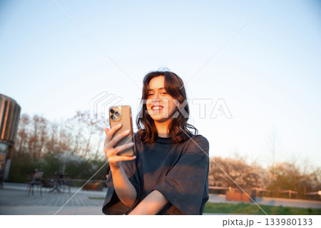 Young woman enjoying a sunset while using her smartphone in a peaceful outdoor setting with blooming trees and clear sky 133980133