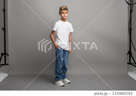 Young boy model with stylish haircut standing in studio. Kid modeling new clothing or showcasing child fashion with copy space on gray background 133980293