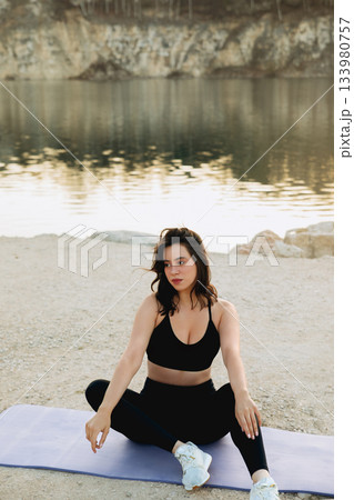 Woman practicing yoga by the calm lake at sunrise, surrounded by nature in a peaceful outdoor setting with rocky cliffs 133980757