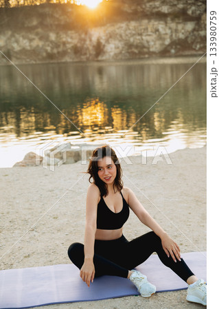 Woman practicing yoga by the calm lake during sunset, enjoying the serene outdoor environment with a peaceful expression Woman practicing yoga by the calm lake during sunset, enjoying the serene outdoor environment with a peaceful expression 133980759