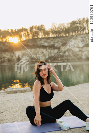 Woman practices yoga at sunrise by a calm lake surrounded by cliffs and trees, focusing on mindfulness and fitness in nature during the early morning 133980761