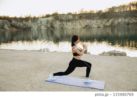 Woman doing lunges on a mat by the calm lake during sunrise near rocky cliffs on a clear morning Woman doing lunges on a mat by the calm lake during sunrise near rocky cliffs on a clear morning 133980769