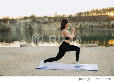 Woman performing lunges on a yoga mat by a tranquil lake during sunset in a natural outdoor setting 133980770