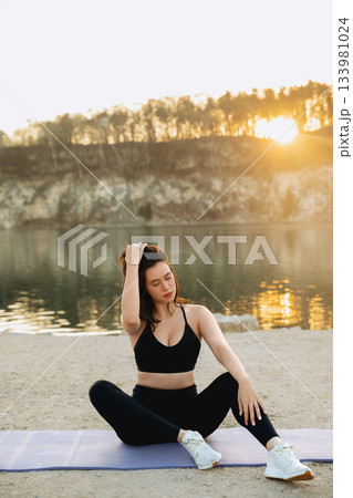 Practicing yoga at sunrise by the lake with warm sunlight reflecting on the water and trees in the background 133981024