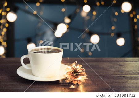 White cup of hot coffee on wooden table with pine cone and warm festive lights. Cozy winter atmosphere, christmas mood, holiday background. 133981712