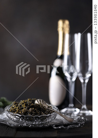Black caviar and champagne. An exquisite snack. Glasses and a bottle of champagne are out of focus. Black caviar is served in a crystal dish with ice. The concept of a festive treat. Black caviar and champagne. An exquisite snack. Glasses and a bottle of champagne are out of focus. Black caviar is served in a crystal dish with ice. The concept of a festive treat. 133981840