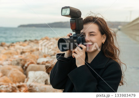 Young female photographer taking pictures outdoors by the sea, smiling while holding a professional camera with flash, capturing moments on a rocky coastline at sunset. 133983479
