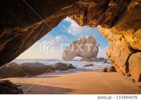 At Praia da Santa Cruz, Penedo do Guincho stands majestically as waves crash into the sandy beach beneath a colorful sunset. 133984076