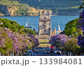 Scenic view of Belem Tower surrounded by blooming jacaranda trees, with sailboats on the Tagus River during sunset in Lisbon, Portugal. 133984081