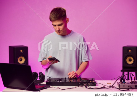 Young DJ with headphones, Skilled DJ engaging with equipment in lively environment, Energetic DJ prepping for live performance amid colorful lighting and fashionable attire 133984487
