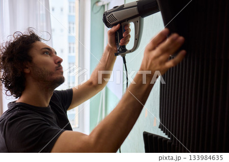 Craftsman carefully detaching acoustic panels indoors, Individual skillfully peels acoustic insulation from wall using heat and precise hand movements for studio setup 133984635