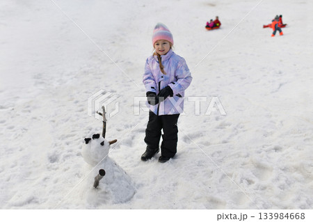 A girl in a hat and gloves makes a snowman in the snow A girl in a hat and gloves makes a snowman in the snow 133984668