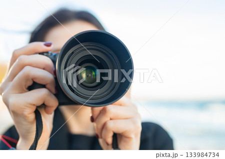 Low-angle view of a female photographer holding a professional camera against the blue sky, capturing outdoor moments and focusing on creative photography. Low-angle view of a female photographer holding a professional camera against the blue sky, capturing outdoor moments and focusing on creative photography. 133984734