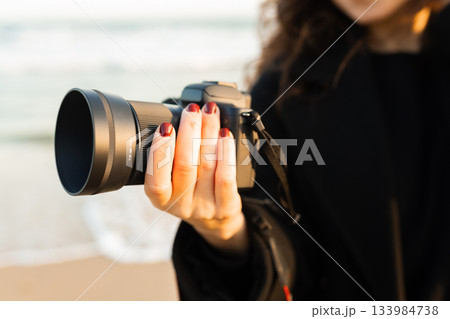 Close-up of woman holding a professional camera on the beach during golden hour, focusing on the lens and capturing outdoor photography moments. Close-up of woman holding a professional camera on the beach during golden hour, focusing on the lens and capturing outdoor photography moments. 133984738