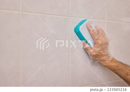 A Man Diligently Cleaning a Tiled Bathroom Wall with His Gloves and a Scrubber Tool A Man Diligently Cleaning a Tiled Bathroom Wall with His Gloves and a Scrubber Tool 133985216