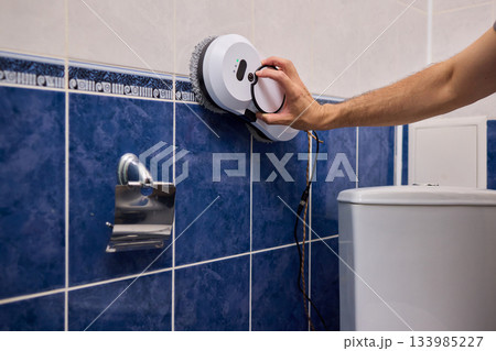 Smart robot performing quick hygienic maintenance in bathroom space, Modern robotic cleaner diligently scrubbing grout lines adjacent to toilet in bright tiled room 133985227