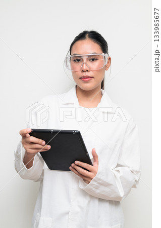 Young Female Scientist in Protective Goggles and Lab Coat on White Background Using Tablet Computer Young Female Scientist in Protective Goggles and Lab Coat on White Background Using Tablet Computer 133985677