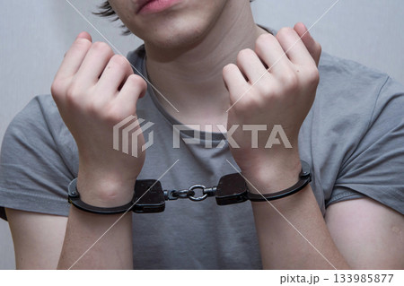 A handcuffed teenager sits on a grey background. Juvenile delinquent, criminal liability of minors. 133985877
