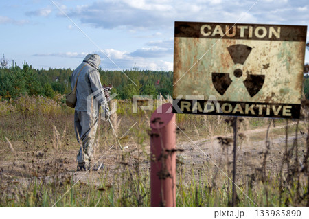Military dosimetrist measures the radiation area, a sign with the inscription: caution, radiation, chemical protective suit and gas mask 133985890