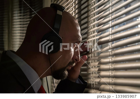 A secret FBI agent Peeps through the blinds and overhears a conversation in headphones, sunlight A secret FBI agent Peeps through the blinds and overhears a conversation in headphones, sunlight 133985907