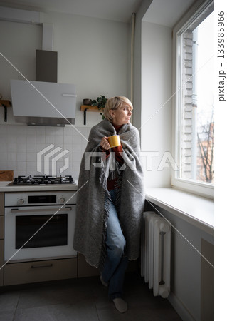 Middle aged woman staying warm in cold unheated apartment wrapping in plaid and drinking hot tea. 133985966