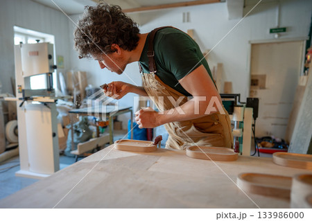Man carpenter blows dust off chisel blade, carefully sanding coasters for glasses with sand paper. Man carpenter blows dust off chisel blade, carefully sanding coasters for glasses with sand paper. 133986000
