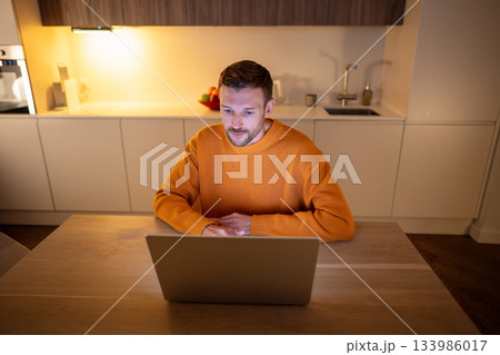Adult businessman freelancer works at home online on laptop, sitting in kitchen late at night. Adult businessman freelancer works at home online on laptop, sitting in kitchen late at night. 133986017