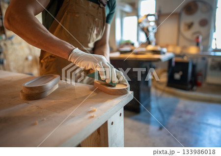 Man carpenter sanding wooden coaster for water glasses, working process in small carpentry workshop. 133986018