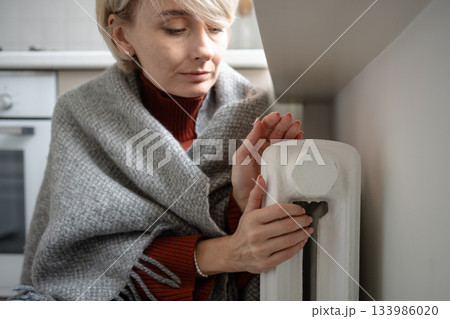 Tired middle aged woman freezing in plaid and warming hands on central heating radiator in apartment 133986020