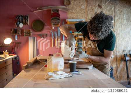 Man carpenter preparing for work on new furniture object, pouring amount of epoxy resin in paper cup 133986021