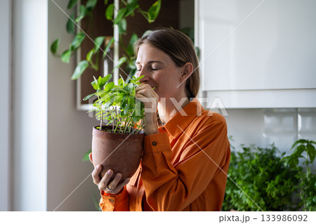 Woman with closed eyes enjoying smelling green mint plant on kitchen at home. Houseplant lover. Woman with closed eyes enjoying smelling green mint plant on kitchen at home. Houseplant lover. 133986092