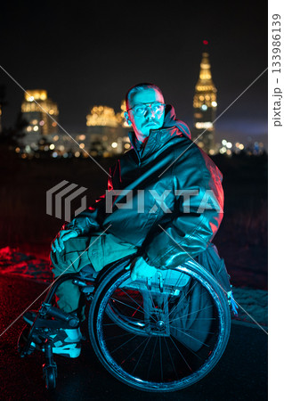 Pondering young disabled man in wheelchair walking outside alone at night in blue neon rgb light. 133986139