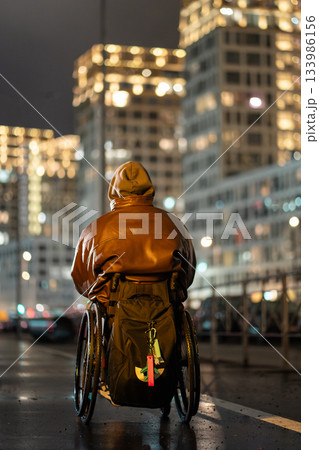Man in wheelchair in hood with backpack walking alone late at night under color rgb light in city. Man in wheelchair in hood with backpack walking alone late at night under color rgb light in city. 133986156